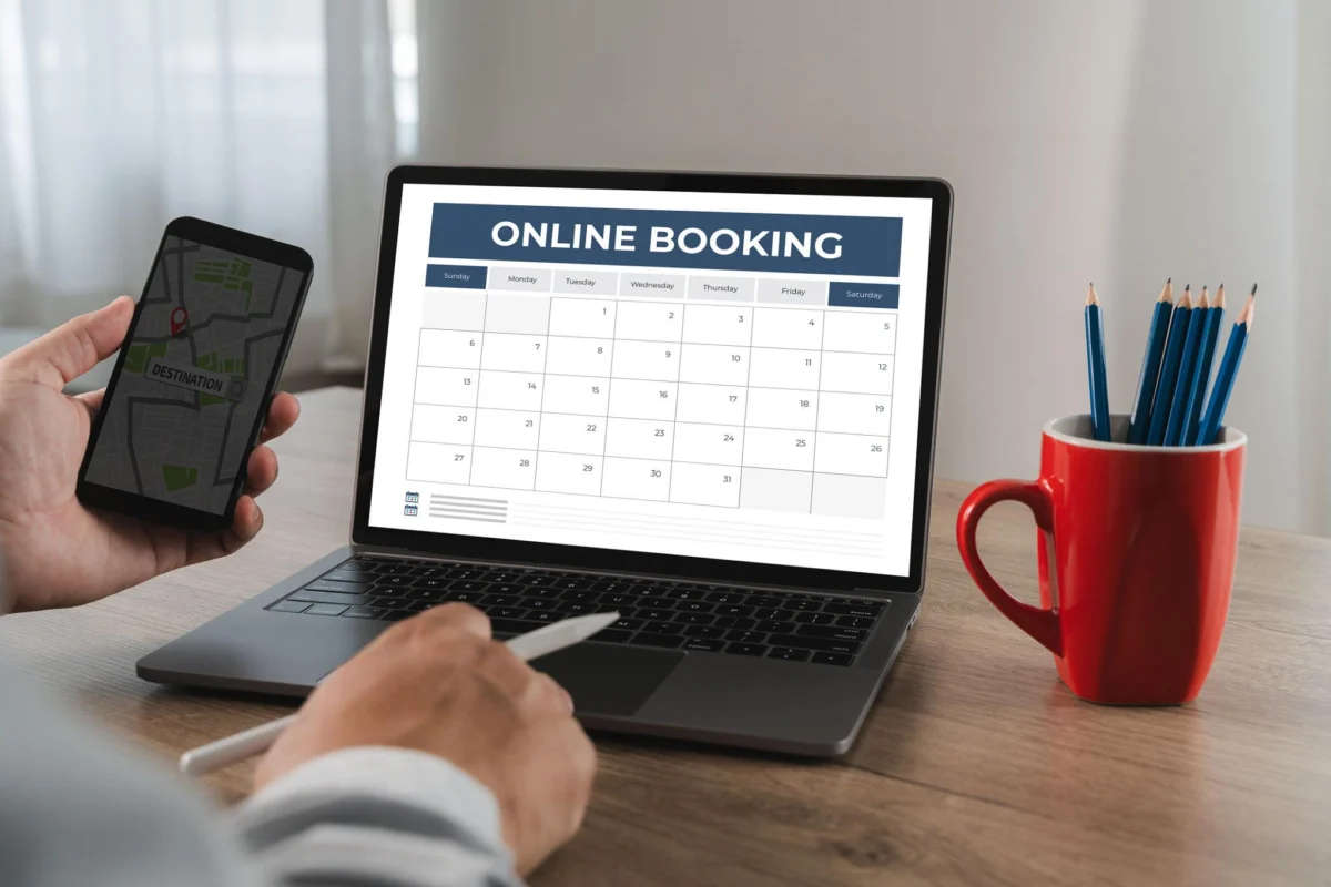 Hands holding a phone beside a laptop showing an online booking calendar
